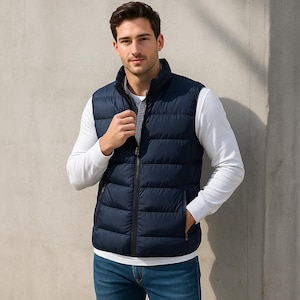 Könnte beinhalten: Eine marineblaue Steppweste mit durchgehendem Reißverschluss und zwei Seitentaschen. Die Weste wird über einem weißen Langarmshirt und einer blauen Jeans getragen. Die Weste hat einen Stehkragen und ein kariertes Innenfutter.