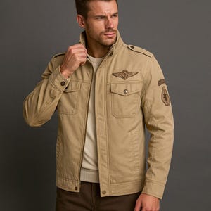 Op de afbeelding: Khaki jas met een opstaande kraag, twee borstzakken en een ritssluiting. De jas heeft een bruin geborduurd embleem op de borst en een embleem op de bovenarm. De jas wordt over een lichtgeleurde trui gedragen.