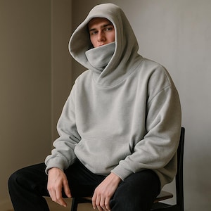 Op de afbeelding: Een lichtgrijze hoodie met een uniek ontwerp met hoge hals en een grote capuchon. De hoodie heeft een kangoeroezak en lange mouwen. De persoon draagt een zwarte broek en zit op een houten stoel.