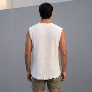 May include: A white sleeveless shirt with a frayed hem and neckline. The shirt is made of a textured fabric. The person is wearing olive green shorts.