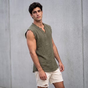 May include: Olive green sleeveless shirt with a V-neck and frayed edges. The shirt is paired with off-white distressed shorts. The model is standing in front of a gray concrete wall. Casual summer wear.