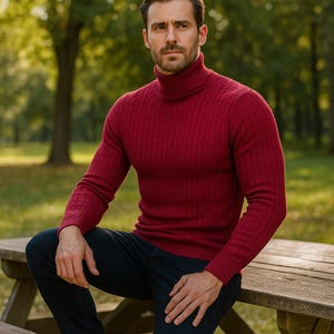 May include: A burgundy cable-knit turtleneck sweater. The sweater has a ribbed texture and a high neckline. The person is wearing dark blue jeans. The background is a park with trees and green grass.
