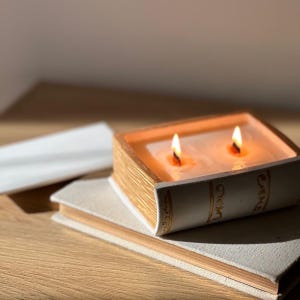 Może przedstawiać: Prostokątna świeca w kształcie książki z białym grzbietem ozdobionym złotymi akcentami. Świeca posiada dwa zapalone knoty w białym wosku, a jej górna krawędź jest teksturowana, aby przypominać strony. Spoczywa na zamkniętej, jasnej książce.