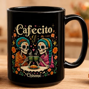Op de afbeelding: Een zwarte keramische mok met een kleurrijke afbeelding van twee skeletten die aan een tafel zitten met koffie en een croissant. De tekst "Cafecito y Chisme" is gedrukt in een decoratief lettertype. Het ontwerp bevat oranje afrikaantjes en sterren.