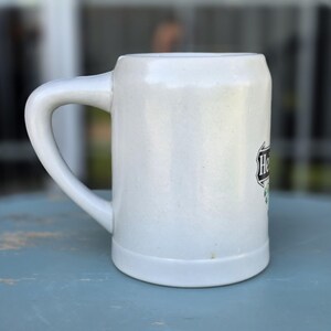 May include: A white ceramic beer stein with a large handle. The stein has a cylindrical shape with a slightly flared base. The word "He" is visible on the side of the stein, along with a green and black design.