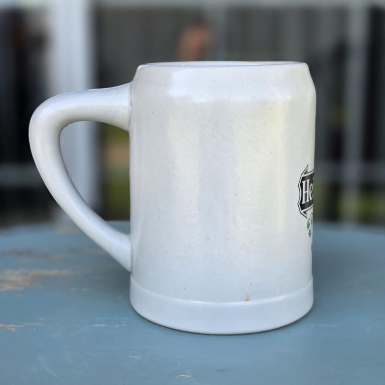 May include: A white ceramic beer stein with a large handle. The stein has a cylindrical shape with a slightly flared base. The word "He" is visible on the side of the stein, along with a green and black design.
