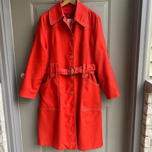 May include: A bright orange trench coat with a matching belt and large collar. The coat features white stitching details and a button-down front. The coat has two front pockets and is hanging on a wooden hanger.