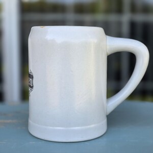 May include: A white ceramic beer stein with a large handle. The stein has a rectangular shape with a slightly textured surface. A black and white emblem is visible on the side of the stein.