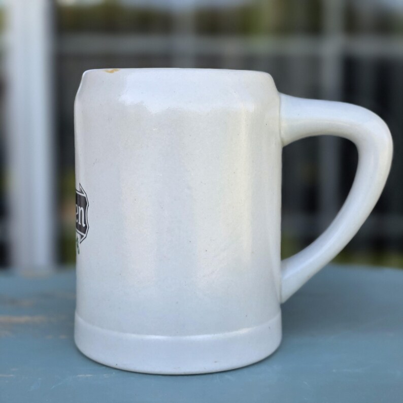 May include: A white ceramic beer stein with a large handle. The stein has a rectangular shape with a slightly textured surface. A black and white emblem is visible on the side of the stein.