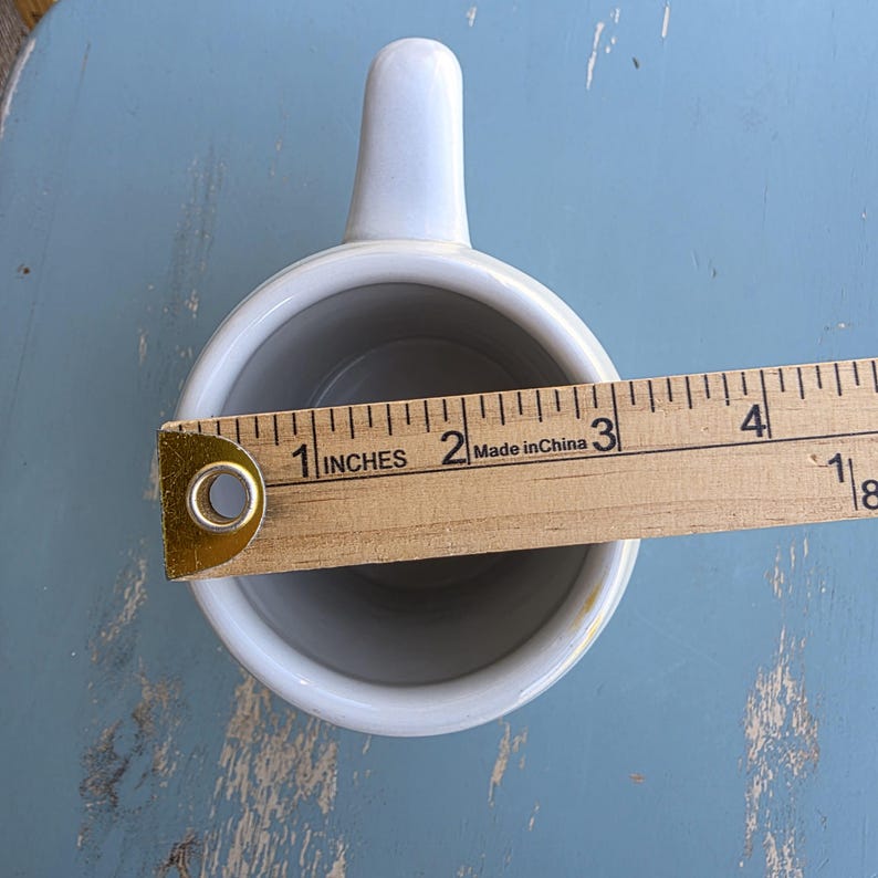 May include: A white ceramic mug with a handle, viewed from above. A wooden ruler is placed across the mug's opening, showing a measurement of approximately 2 inches. The mug's interior is gray.