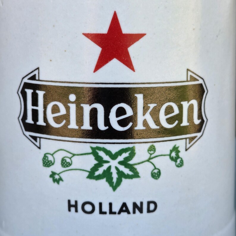 May include: Close-up of a white can with the Heineken logo. The logo features a red star at the top, the word "Heineken" in white on a black and gold banner, and a green leaf design. The word "HOLLAND" is printed at the bottom.