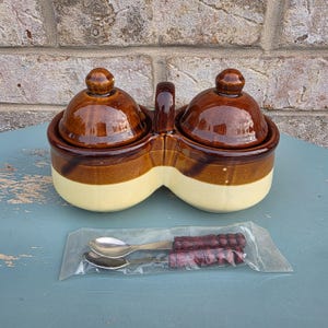 May include: A double-chambered ceramic condiment server featuring two round containers with matching lids, finished in a two-tone brown and cream glaze. A central handle connects the two bowls. Two small metal spoons with textured brown handles are included in a clear plastic sleeve.