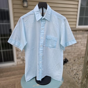 Puede incluir: Una camisa de manga corta de color azul claro con un sutil estampado de lunares blancos, cuello en punta y un bolsillo en el pecho en el lado izquierdo. La camisa se muestra en una percha negra y tiene una etiqueta de marca visible en el cuello.