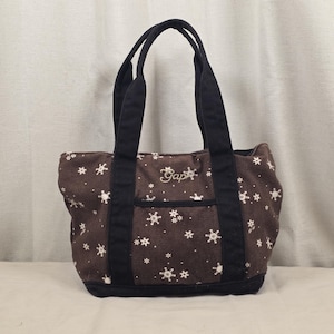 May include: Brown tote bag with black handles and trim. The bag features a white snowflake pattern and the embroidered word "Gap". The tote has a small front pocket and is made of a sturdy fabric.