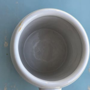 May include: Overhead view of a white ceramic mug with a rounded handle. The interior is a light gray, and the exterior is a matte white. A small yellow stain is visible on the rim. The mug sits on a light blue surface.