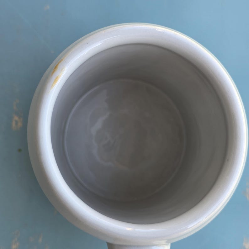 May include: Overhead view of a white ceramic mug with a rounded handle. The interior is a light gray, and the exterior is a matte white. A small yellow stain is visible on the rim. The mug sits on a light blue surface.