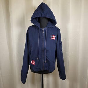 May include: Navy blue hooded zip-up jacket with American flag patches. The jacket has a drawstring hood, a full-length zip, and a small pocket on the left sleeve. The American flag patches are red, white, and blue.