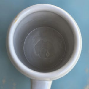 May include: Overhead view of a white ceramic mug with a handle. The interior of the mug is gray, and the rim is white. A small yellow stain is visible on the rim. The mug is set against a light blue background.