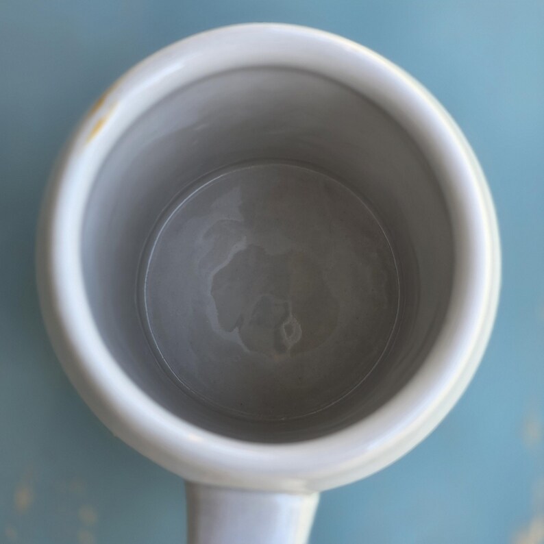 May include: Overhead view of a white ceramic mug with a handle. The interior of the mug is gray, and the rim is white. A small yellow stain is visible on the rim. The mug is set against a light blue background.