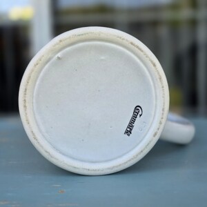 May include: A white ceramic mug base with a slightly textured rim. The bottom of the mug has the word "Ceramarte" printed in black script. The mug is set against a blurred background.