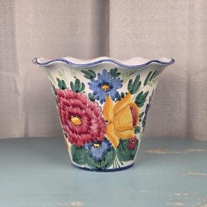 Op de afbeelding: Een witte keramische bloempot met een geschulpte rand en een blauwe rand. De pot is versierd met handgeschilderde bloemen in roze, geel en blauw, met groene bladeren. De pot staat op een lichtblauwe ondergrond.