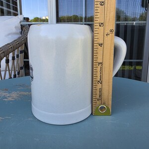 May include: A white ceramic beer stein with a handle. The stein has a dark emblem on the front. A wooden ruler is next to the stein, measuring approximately 6 inches in height.