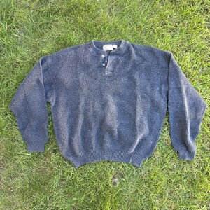 May include: A dark charcoal gray long-sleeve sweater featuring a textured waffle knit pattern, a ribbed crew neckline, and a partial button-down placket with two visible buttons at the center front. The sweater has ribbed cuffs and a ribbed hem.