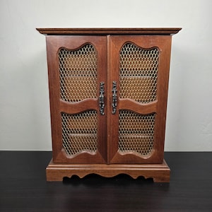 Op de afbeelding: Een houten sieradenkast met twee deuren. De deuren hebben een decoratief gaasontwerp en sierlijke metalen handgrepen. De kast is warmbruin van kleur en staat op een bijpassende basis. De kast heeft een klassieke, vintage uitstraling.