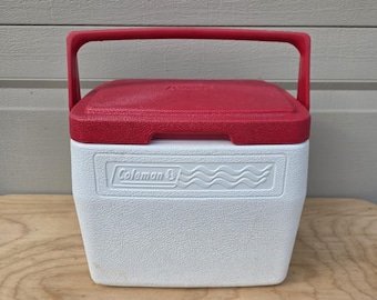 Puede incluir: Una nevera portátil rectangular Coleman con base blanca y tapa roja. La nevera cuenta con un asa roja abatible y un logotipo moldeado en la parte frontal que dice Coleman junto a un diseño de ondas. El exterior tiene un acabado texturizado.