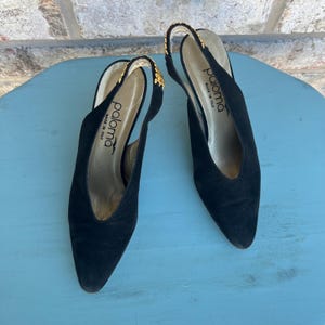 Puede incluir: Un par de zapatos de tacón Paloma negros con punta. Los tacones tienen detalles dorados en las correas y el nombre de la marca es visible en la plantilla. Los zapatos están hechos en Italia.