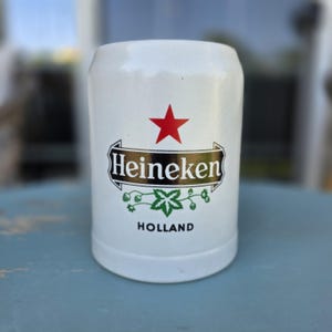 May include: White ceramic beer stein with the Heineken logo, featuring a red star, the brand name, and a green hop leaf design. The word "HOLLAND" is printed below the logo. The stein has a cylindrical shape.