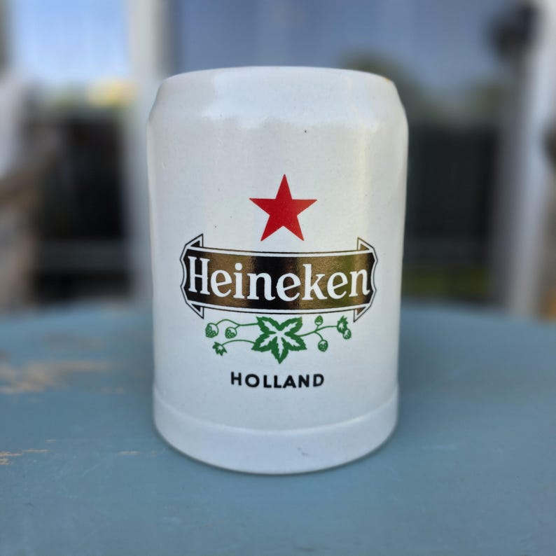 May include: White ceramic beer stein with the Heineken logo, featuring a red star, the brand name, and a green hop leaf design. The word "HOLLAND" is printed below the logo. The stein has a cylindrical shape.