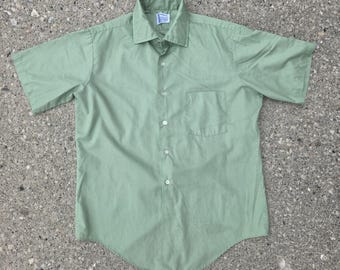 May include: A short-sleeved button-down shirt in a solid sage green colour. The shirt features a pointed collar, a single chest pocket on the left side, and a front placket with five visible white buttons. The hem is curved at the bottom.