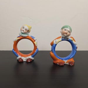 May include: Two vintage ceramic napkin rings. Each ring features a small, detailed figure of a child holding a colorful ring. One child wears a yellow hat, the other a green hat. The rings are orange and blue.