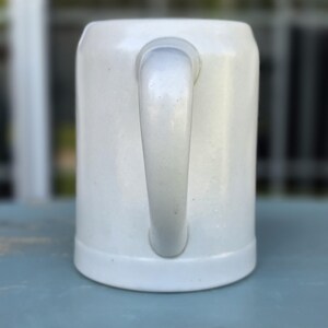 May include: A white ceramic beer stein with a curved handle. The stein has a cylindrical shape with a slightly flared base. The handle is attached to the side of the stein. The background is blurred, suggesting an outdoor setting.