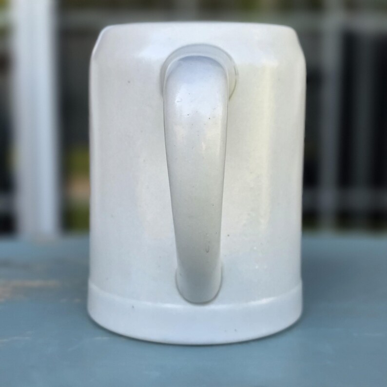 May include: A white ceramic beer stein with a curved handle. The stein has a cylindrical shape with a slightly flared base. The handle is attached to the side of the stein. The background is blurred, suggesting an outdoor setting.