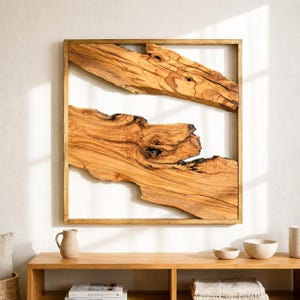 May include: A square wall art piece featuring a thin wooden frame enclosing two horizontal, irregular slabs of light brown wood with visible grain patterns and dark knots, separated by a clear space against a plain white background.