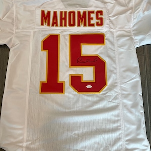 May include: White football jersey with red and gold accents. The jersey features the name "MAHOMES" across the shoulders and the number "15" on the front and back. The jersey is signed and has a small authentication sticker.