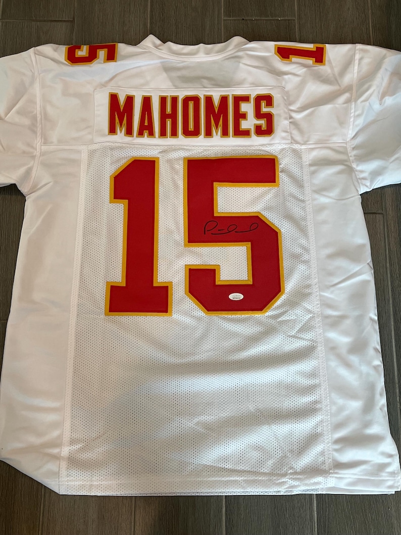 May include: White football jersey with red and gold accents. The jersey features the name "MAHOMES" across the shoulders and the number "15" on the front and back. The jersey is signed and has a small authentication sticker.