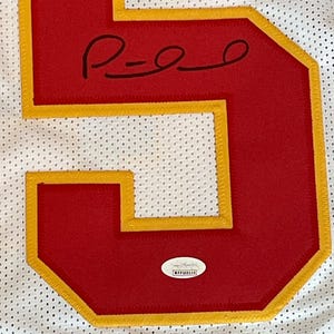 May include: Close-up of a white jersey with a large red number 5 outlined in gold. The number has a signature in black ink. A small, oval authentication sticker is at the bottom.