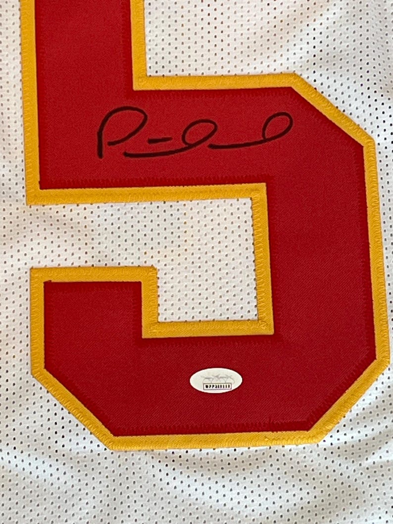 May include: Close-up of a white jersey with a large red number 5 outlined in gold. The number has a signature in black ink. A small, oval authentication sticker is at the bottom.