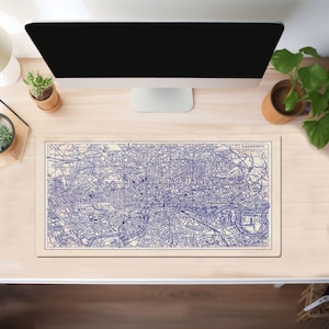 以下が含まれることがあります： ロンドンマップが印刷された青と白のデスクマット。地図には街の通りとランドマークが示されています。マットは、コンピューターモニターと植物のある木製デスクの上に置かれています。