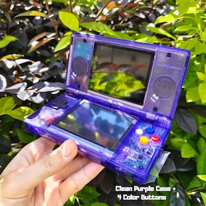 May include: A handheld video game console with a transparent purple plastic case, featuring two screens, a directional pad, and four colored action buttons. The text Clean Purple Case + 4 Color Buttons is printed in the bottom right corner.