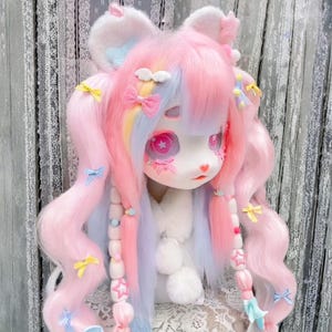 Può includere: Una testa di bambola con capelli rosa pastello e blu acconciati a trecce, decorata con fiocchi e accessori a forma di stella. La bambola ha grandi occhi espressivi e indossa una soffice sciarpa bianca. L'estetica generale è morbida e stravagante.