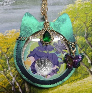 Op de afbeelding: Een groen katvormig accessoire met een gouden ketting en een groene edelsteen hanger. Het accessoire heeft een cirkelvormige afbeelding met een stripfiguur en een kleurrijke vlinderbedel.