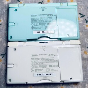 Puede incluir: Dos consolas de juegos port&aacute;tiles Nintendo DS Lite. Una es azul claro, la otra es blanca. Se ven las partes traseras de las consolas, mostrando informaci&oacute;n del modelo, n&uacute;meros de serie y detalles de la bater&iacute;a. El texto "NINTENDO DS Lite" est&aacute; impreso en ambas.