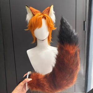 Può includere: Un set cosplay da volpe con orecchie arancioni con dettagli in pelliccia bianca, una parrucca arancione e una lunga coda soffice con una sfumatura di nero, arancione e marrone. La coda è tenuta da una mano.
