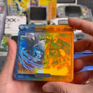 Puede incluir: Una funda de cartucho de juego portátil, dividida en secciones azules y naranjas. La funda presenta el logotipo de "Pokemon" e imágenes de dos personajes Pokémon. La funda está sujeta en la mano de una persona.