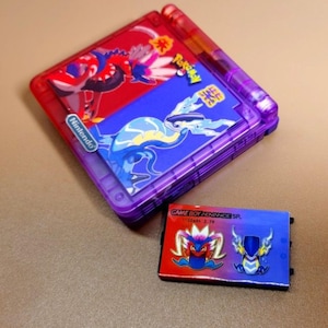 Puede incluir: Una consola Game Boy Advance SP roja y morada con un cartucho a juego. La consola presenta ilustraciones de Pokémon y un logotipo de Nintendo. El cartucho tiene un diseño rojo y azul con personajes de Pokémon.