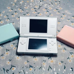 Puede incluir: Una consola de juegos port&aacute;til Nintendo DS blanca est&aacute; abierta, mostrando dos pantallas negras. Tambi&eacute;n se ven dos consolas adicionales, una azul claro y otra rosa. Las consolas est&aacute;n sobre una superficie con estampado floral.
